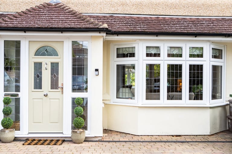 Front exterior of a cream-colored house with cream door, matching windows, and brick driveway with potted plants