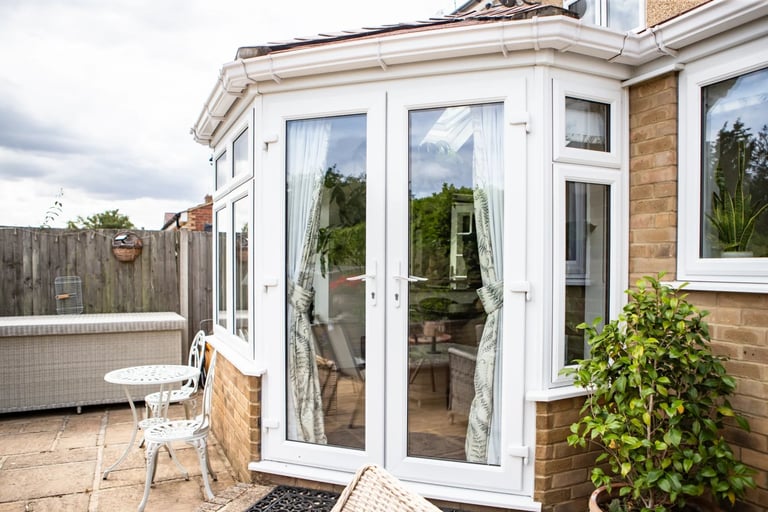 Modern white uPVC conservatory with glass doors attached to brick house, featuring potted plants and garden seating area