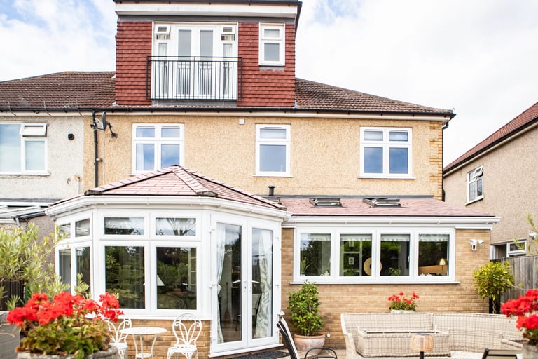 Residential house with modern white glass conservatory extension, red-tiled mansard roof, and landscaped garden with red flowers