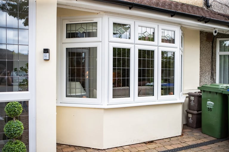 Modern white bay window with multiple glass panes on a cream-colored house exterior, brick patio visible with green waste bin