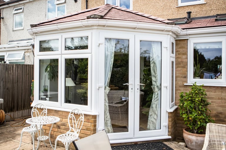 Modern white conservatory extension with glass doors and windows attached to a brick house, featuring white patio furniture outside