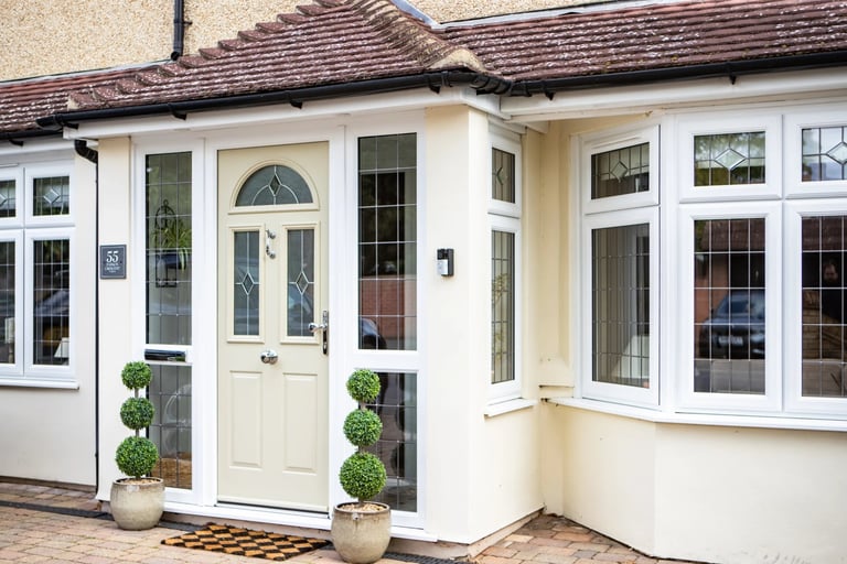 Modern white conservatory extension with glass doors and windows, cream-colored walls, tiled roof, and potted topiaries flanking the entrance door